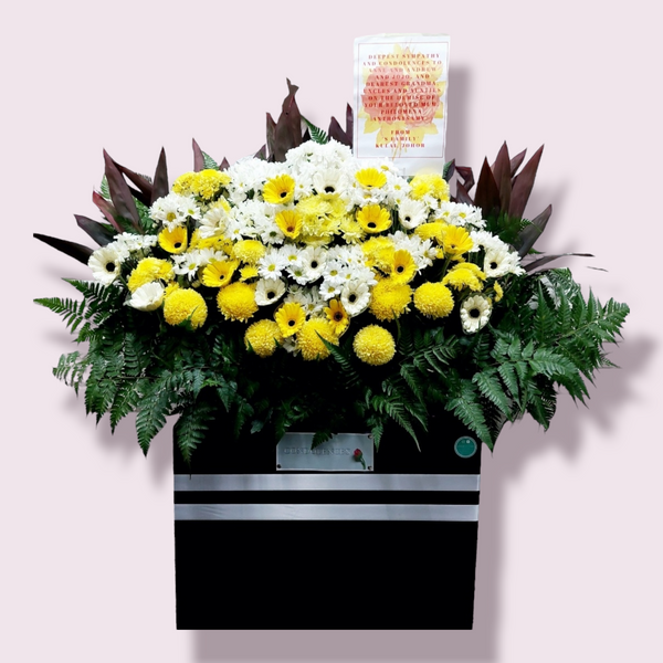 A floral condolence wreath arrangement with mainly white and yellow flowers, ferns, and leaves in a box stand, accompanied by a laminated A4 sized card with a message.