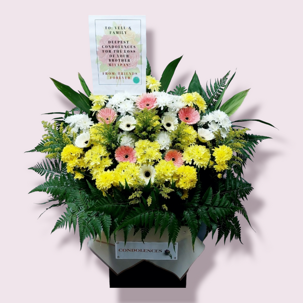 A floral condolence wreath arrangement with mainly white and yellow flowers, ferns, and leaves in a box stand, accompanied by a laminated A4 sized card with a message.